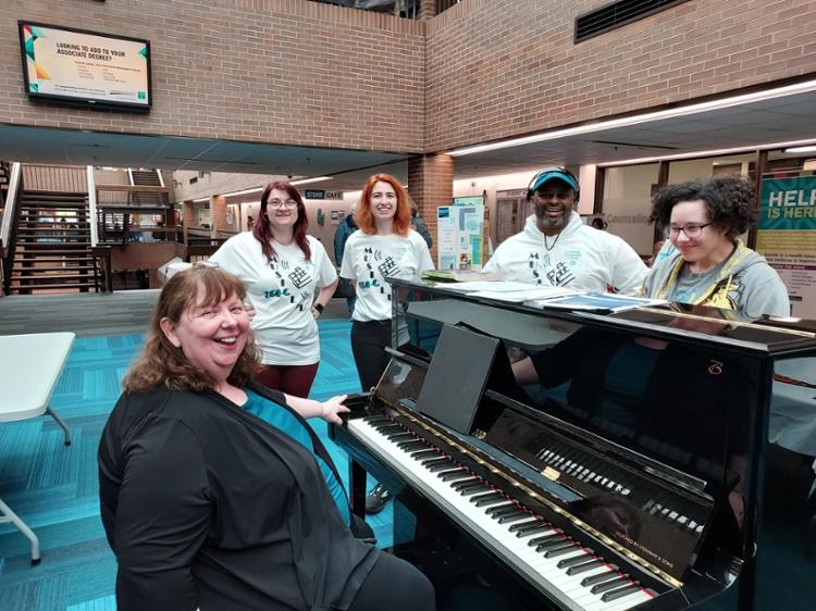 Tri-C West Campus Music Club members gathered around the piano in matching tee shirts