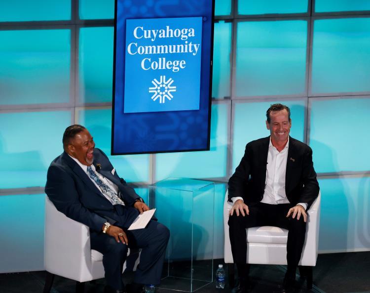 Two men in business attire sitting on stage in conversation, smiling