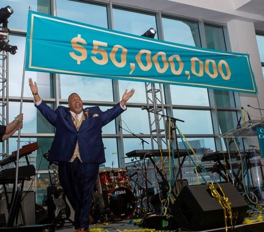 Dr. Michael Baston, arms raised, on stage in front of banner announcing $50 million goal achieved
