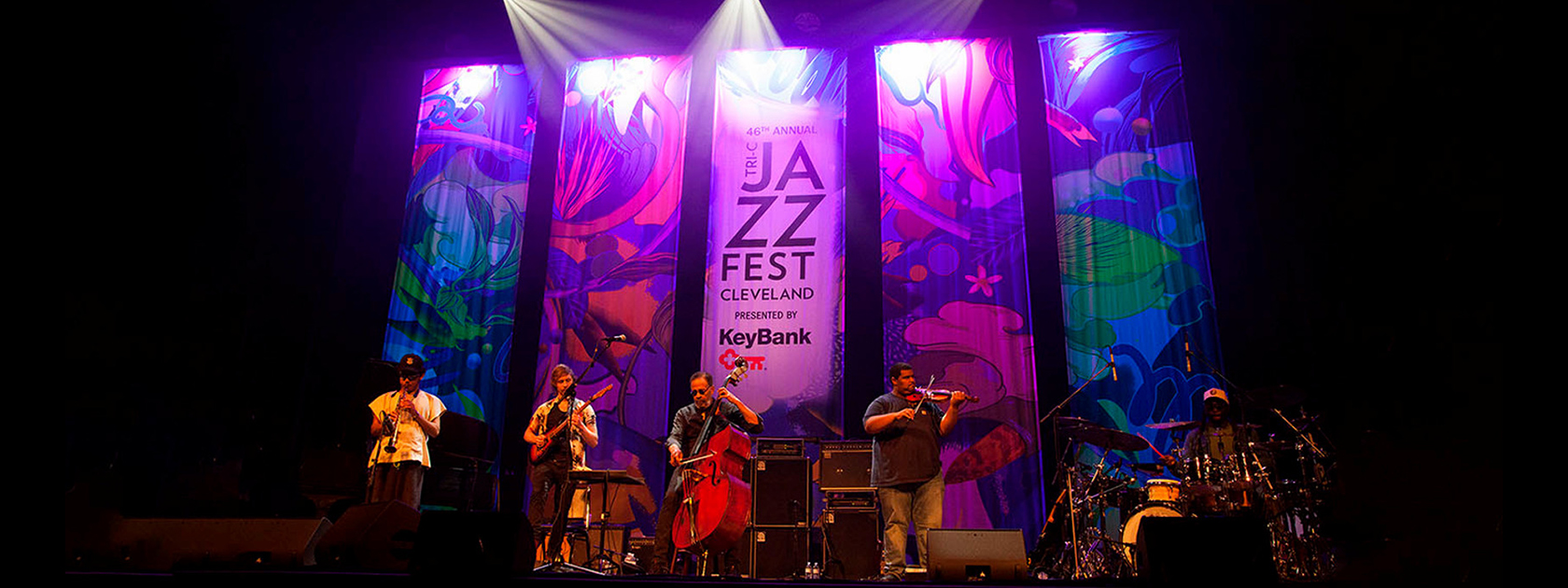 View of a stage with a colorful backdrop that says "Tri-C JazzFest Cleveland presented by KeyBank." There are five musicians on stage with instruments.