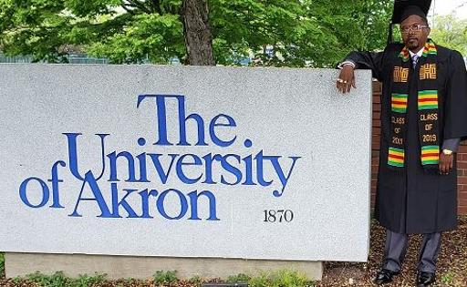 Darcious Linton Sr. wearing a cap and gown while standing next to a University of Akron sign