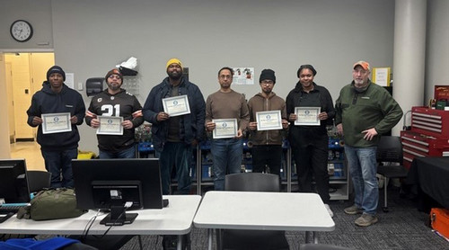 A group of Fiber Optic students stands together in a classroom or training space, holding their Certificates of Completion earned for successfully completing the program. The students appear proud and confident as they pose for a group photo following the completion of hands-on fiber optic training
